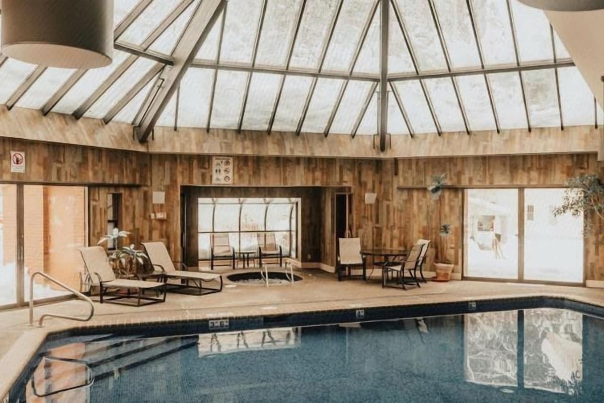 An indoor swimming pool with lounge chairs, a glass ceiling, and wooden walls.