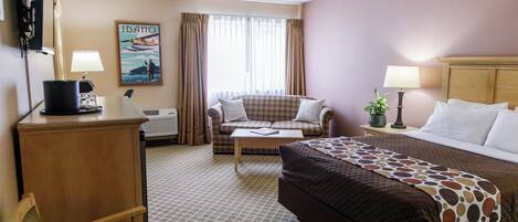 A hotel room with a bed, a sofa, a desk, a chair, a lamp, a plant, and a painting on the wall.
