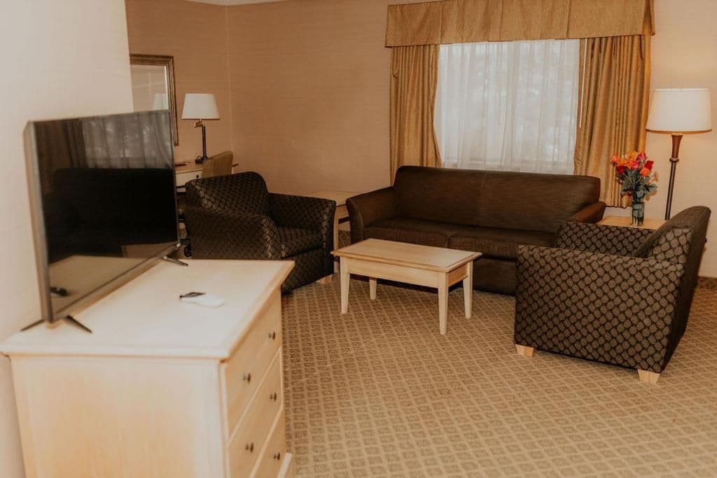 A hotel room with a TV, a sofa, two patterned armchairs, a small table, and a dresser.