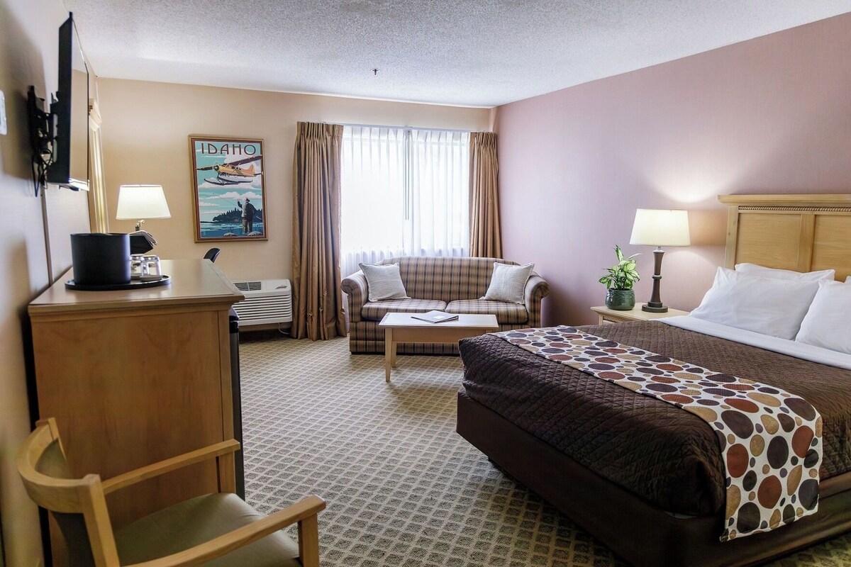 A hotel room with a bed, a sofa, a desk with a lamp, a chair, a window with curtains, and a painting on the wall.