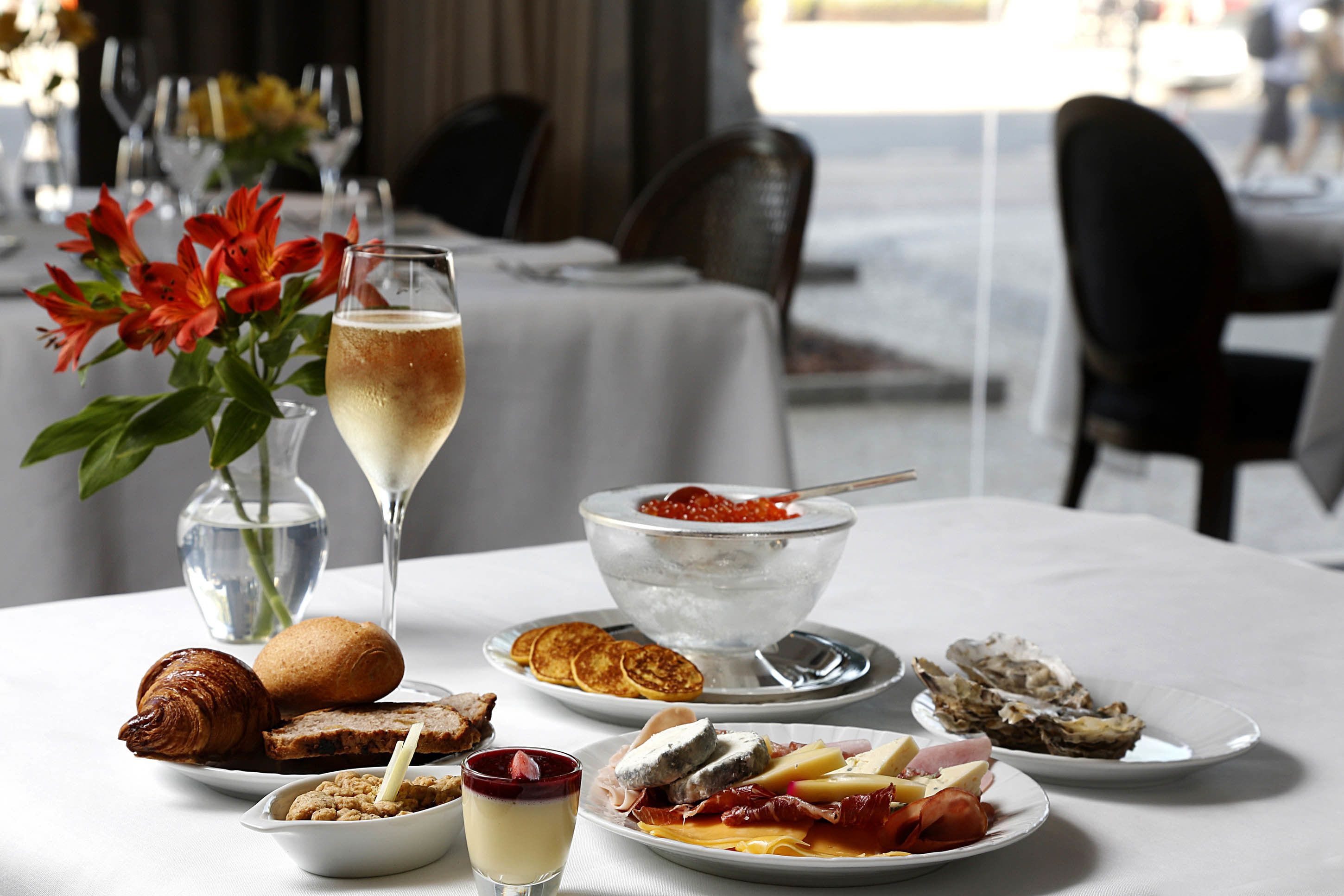 Uma mesa de jantar com diversos pratos, incluindo um croissant, queijo e ostras, além de uma taça de champanhe.