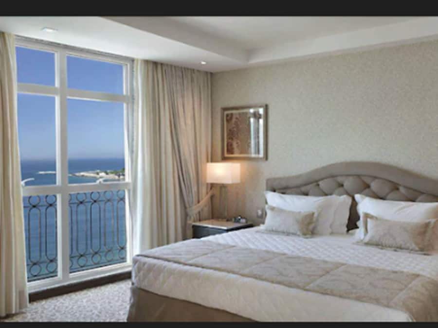 A hotel room with a large window, a bed with multiple pillows, a nightstand, and a framed picture on the wall.