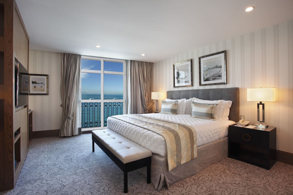 Habitación de hotel con una cama grande, un banco, una mesita de noche y vista al océano desde un balcón.