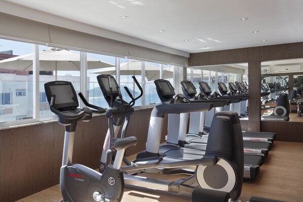 Un gimnasio con varias cintas de correr y bicicletas estáticas, ventanas amplias y vistas a edificios exteriores.