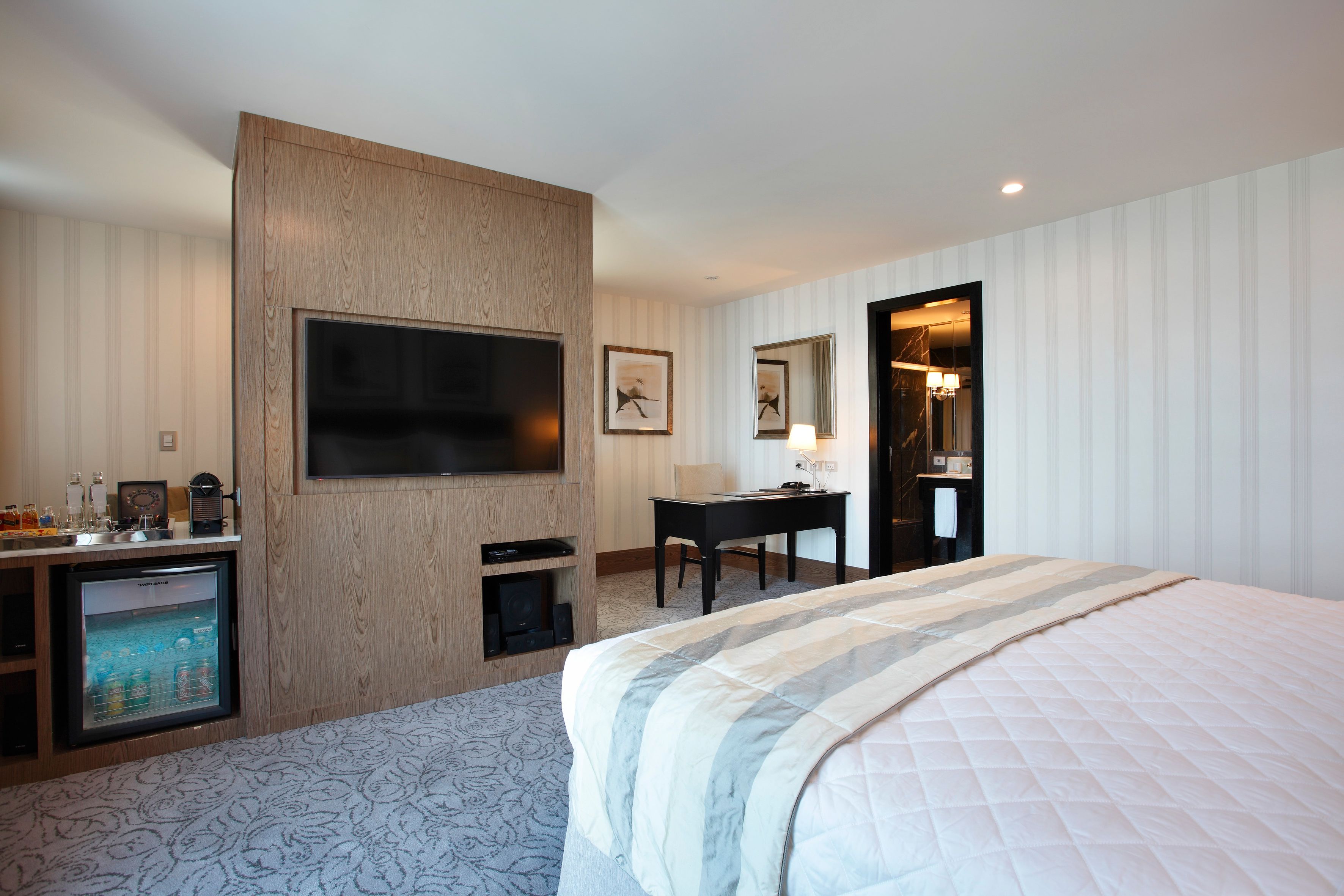 Quarto de hotel com uma cama grande, televisão de tela plana presa a um painel de madeira, escrivaninha com uma cadeira e uma pequena geladeira.