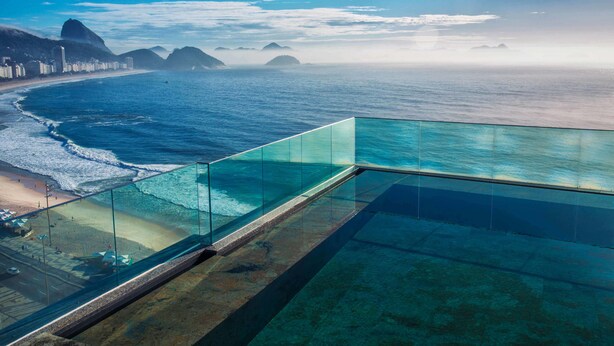 Una piscina con fondo de cristal que ofrece vistas infinitas a una playa con aguas azules cristalinas y montañas a lo lejos.