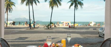 A table set with various dishes and drinks, overlooking a beach with palm trees and people in the background.