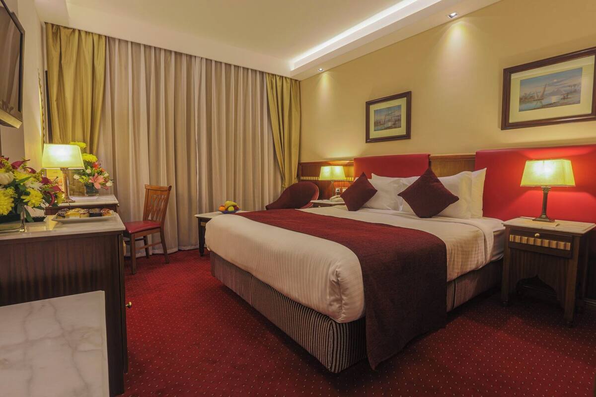 Habitación de hotel con una cama grande, dos mesitas de noche con lámparas, un escritorio con lámpara y flores, una silla y una ventana con cortinas.
