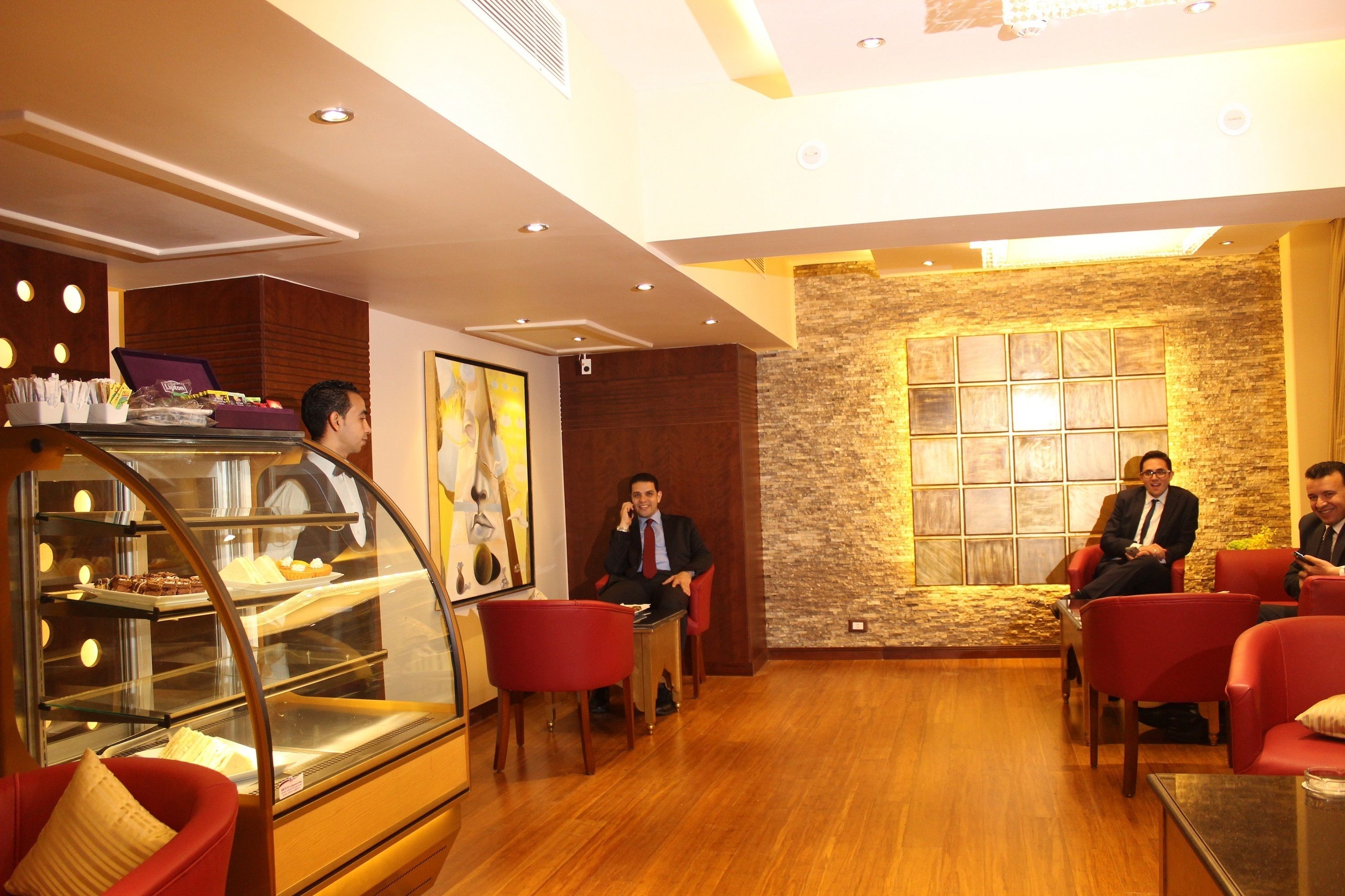 A modern hotel lobby with a bakery display, wooden flooring, and red seating.