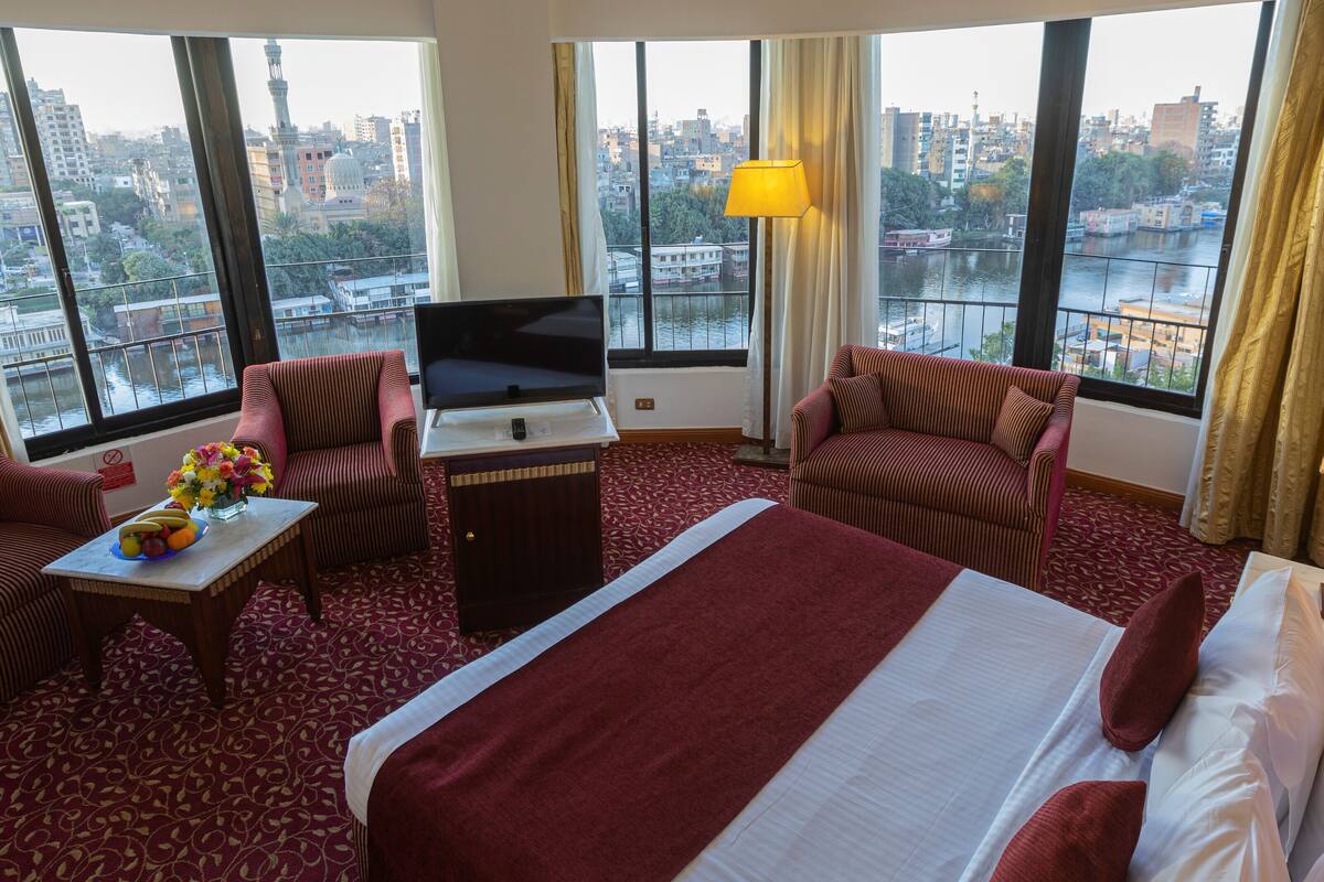 Habitación de hotel con una cama grande, dos sillones, una mesita con un jarrón de flores, un televisor y vistas a un paisaje urbano a través de amplios ventanales.