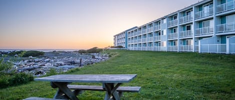 A beachfront hotel with multiple balconies, a grassy lawn, and a wooden picnic table.