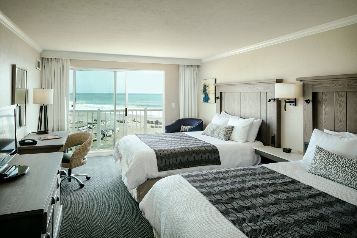 A hotel room with two beds, a desk, a chair, and a large window overlooking the beach.