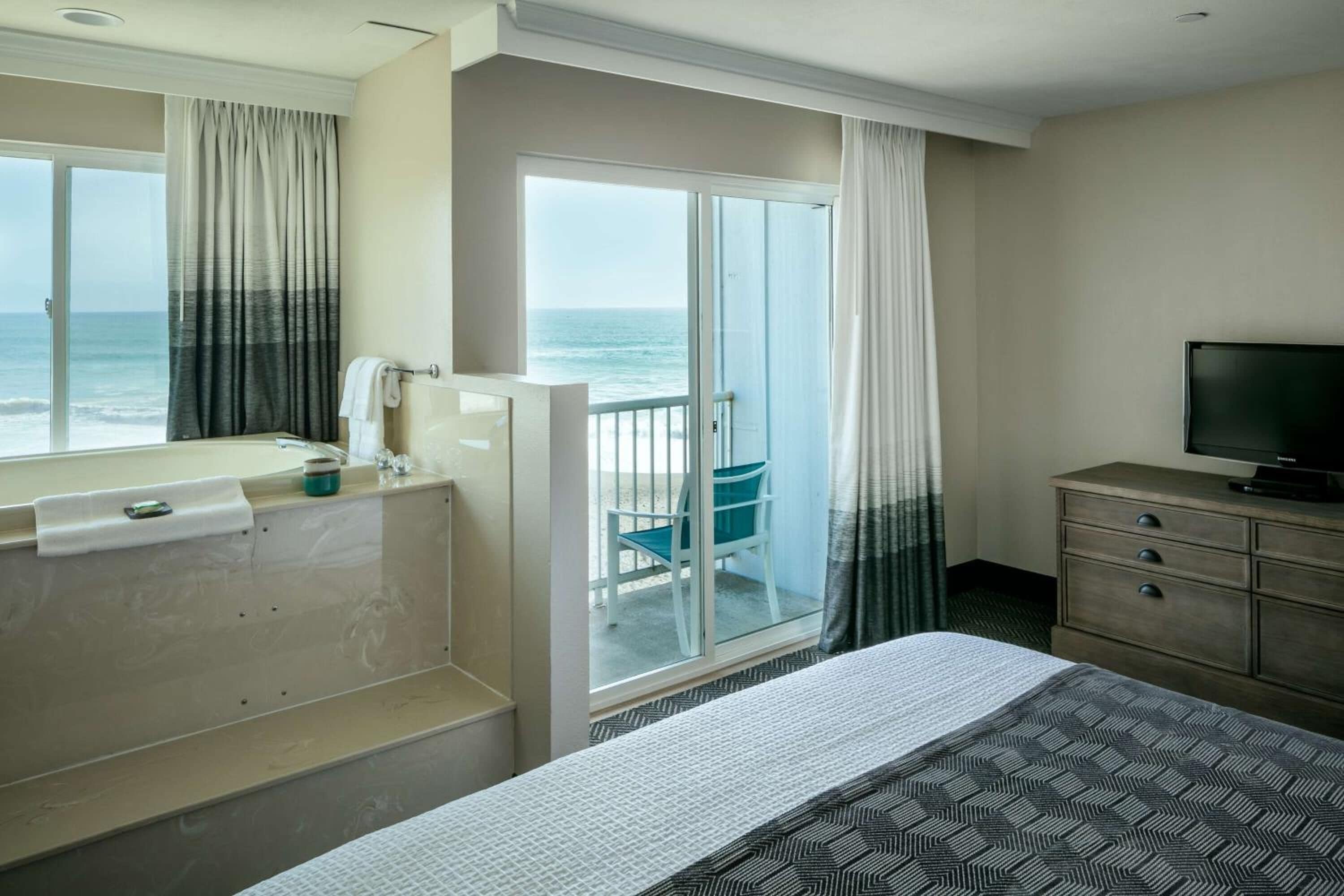 A hotel room with a large bed, a television, a bathroom with a bathtub, and a view of the ocean through the sliding glass door.