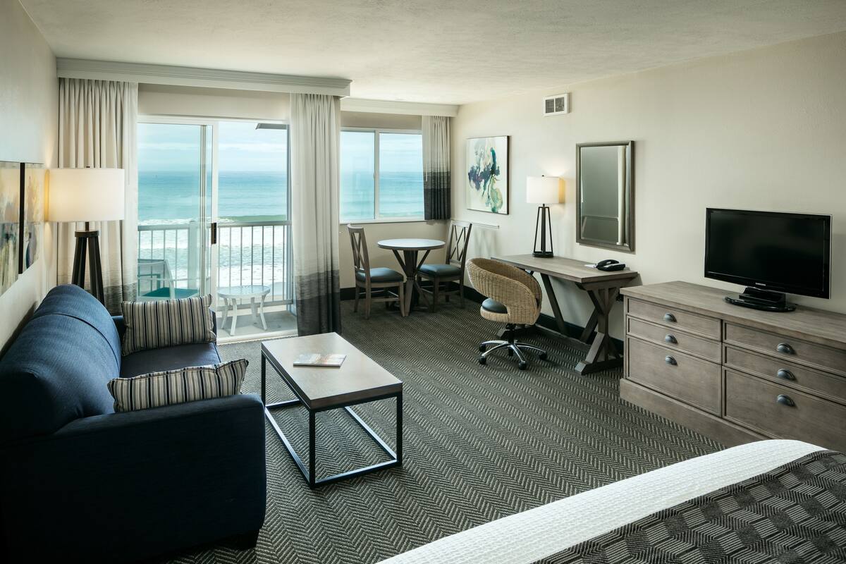 A hotel room with a bed, a sofa, a desk, a TV, a chair, a table, and a view of the ocean.