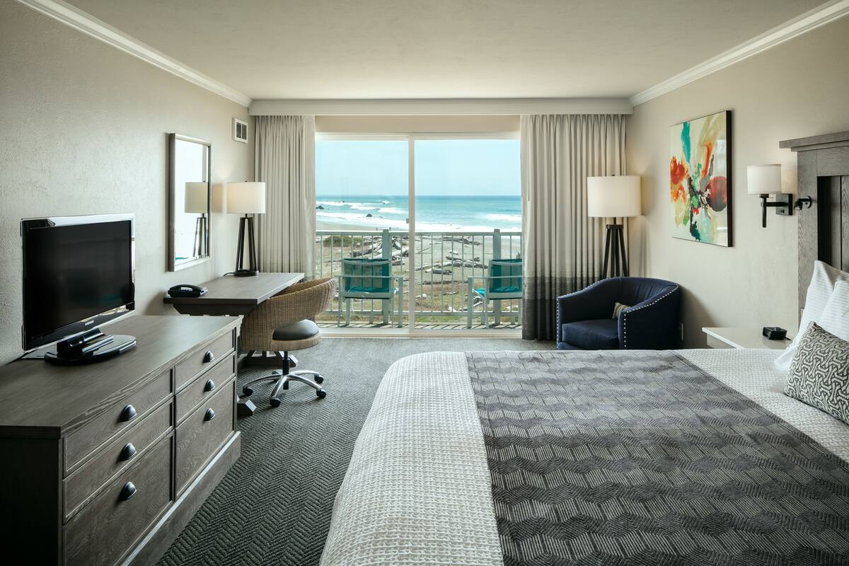A hotel room with a bed, a desk, a TV, a chair, a balcony with a view of the beach, and a painting on the wall.