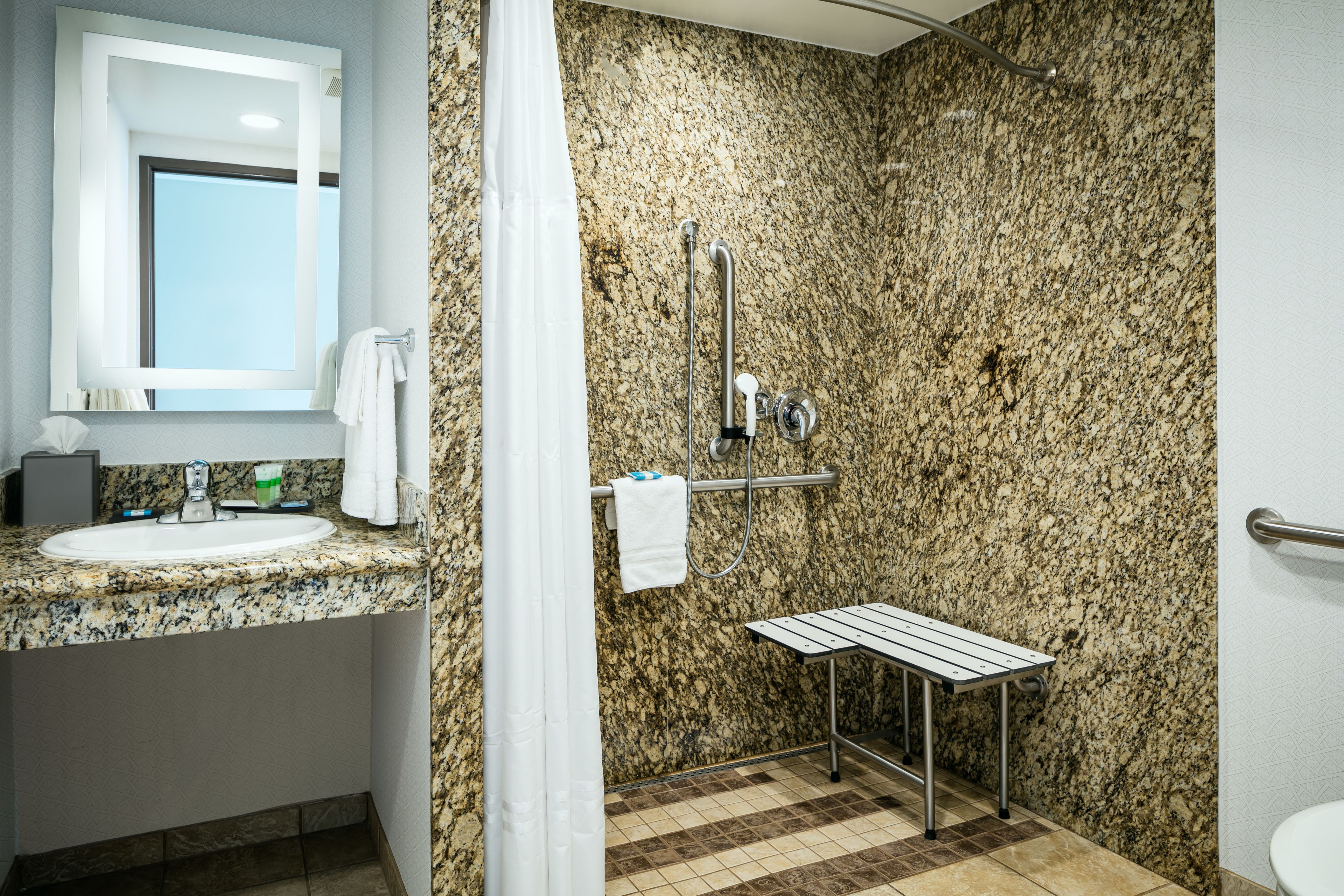 A bathroom with a granite countertop sink, a mirror, a shower with a grab bar, and a toilet.