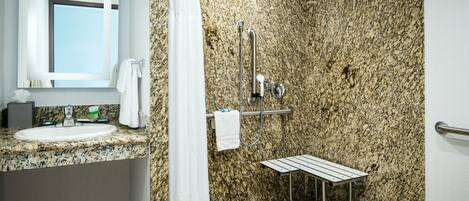 A bathroom with a granite countertop sink, a mirror, a shower with a grab bar, and a toilet.