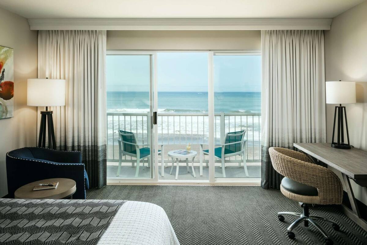 A hotel room with a large sliding glass door leading to a balcony with a view of the ocean. There are two chairs and a small table on the balcony.
