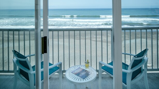 A balcony with a view of the ocean, two chairs, a table with a magazine, and a bottle of water. - Beachfront Inn (Brookings)