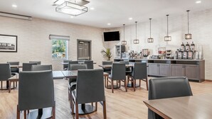 A modern hotel dining area with wooden tables and chairs, a kitchen counter with coffee makers, and a large window with a view of greenery. - Beachfront Inn (Brookings)