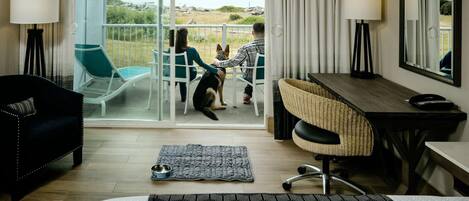 Ein Hotelzimmer mit Bett, Schreibtisch, Stuhl und einem Balkon mit Blick auf den Strand, auf dem ein Hund sitzt.