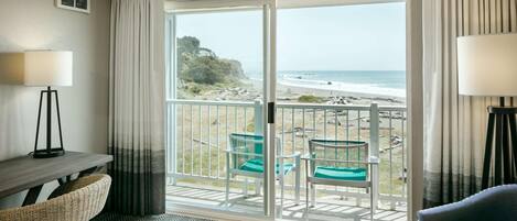 A hotel room with a balcony overlooking a beach, featuring a desk, chairs, and a view of the ocean.