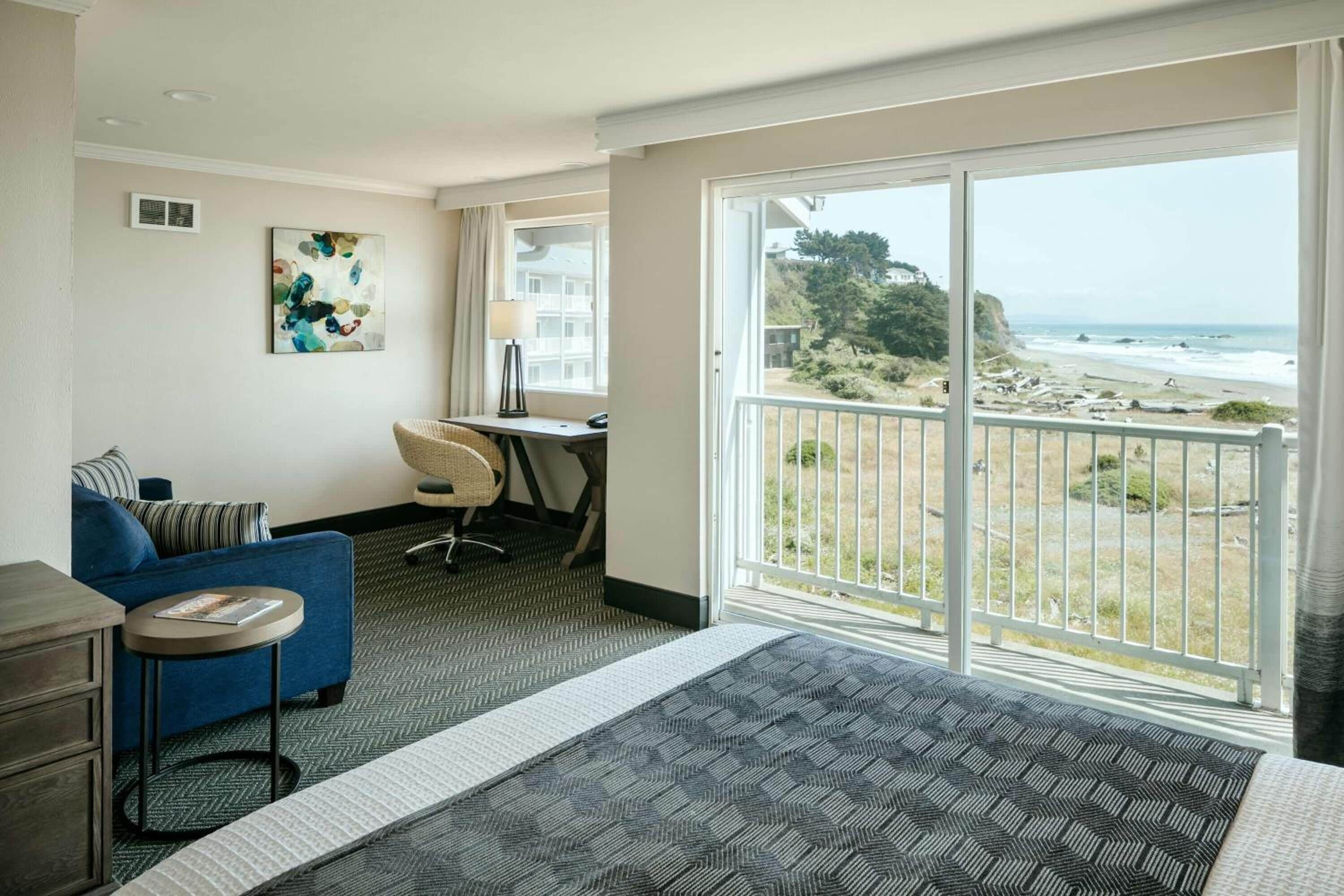 A hotel room with a bed, a desk, a chair, a sofa, and a large window with a beach view.