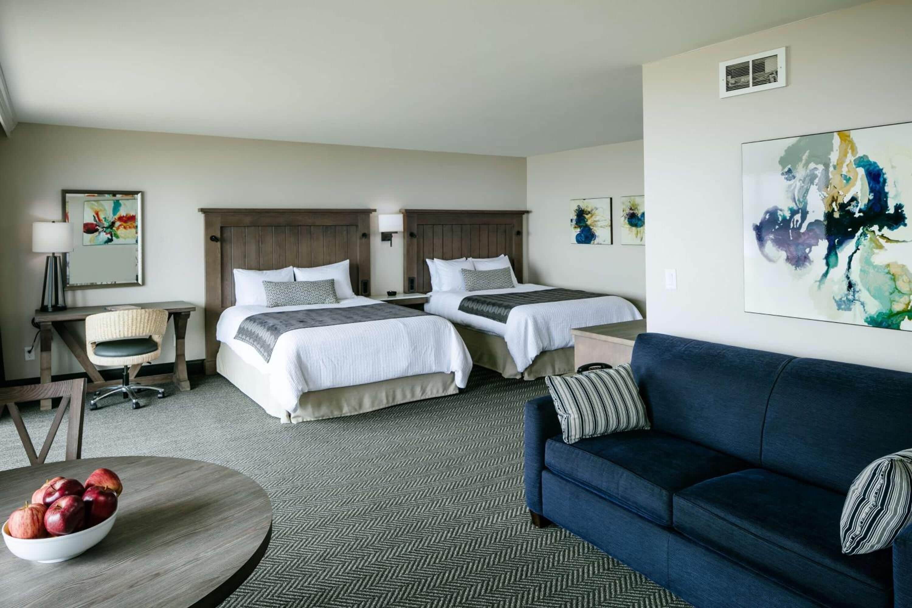 A hotel room with two beds, a blue sofa, a round table with a bowl of fruit, and artwork on the walls.