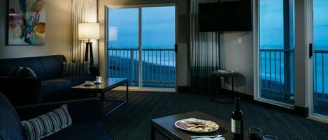 Habitación de hotel con balcón, un sofá, una mesa con comida y vino, y un televisor montado en la pared.