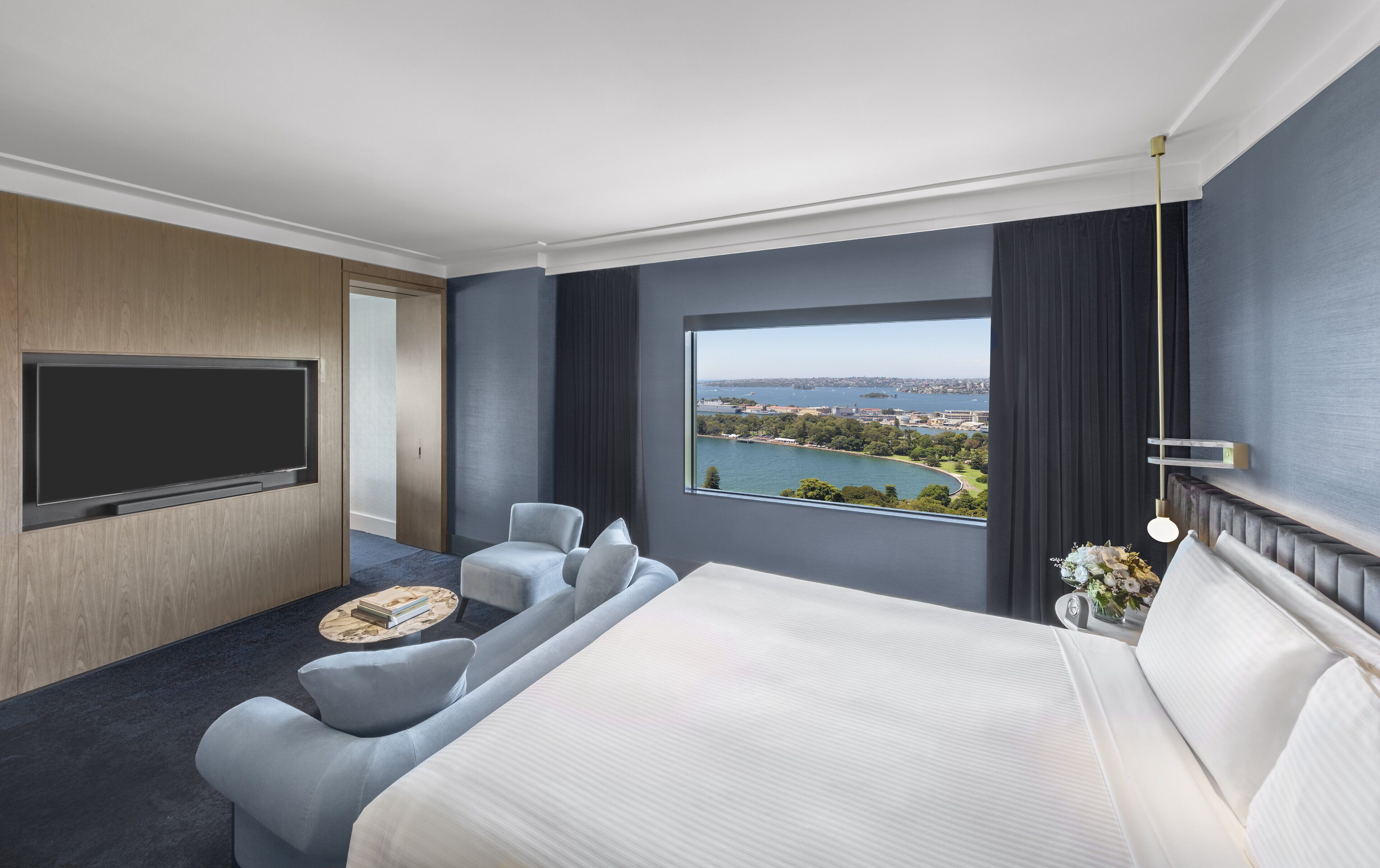 Habitación de hotel con una cama grande, un televisor montado en la pared, una zona de estar con una pequeña mesa y una ventana que ofrece vistas a una masa de agua y un horizonte urbano distante.