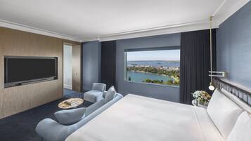 Habitación de hotel con una cama grande, un televisor montado en la pared, una zona de estar con una pequeña mesa y una ventana que ofrece vistas a una masa de agua y un horizonte urbano distante.