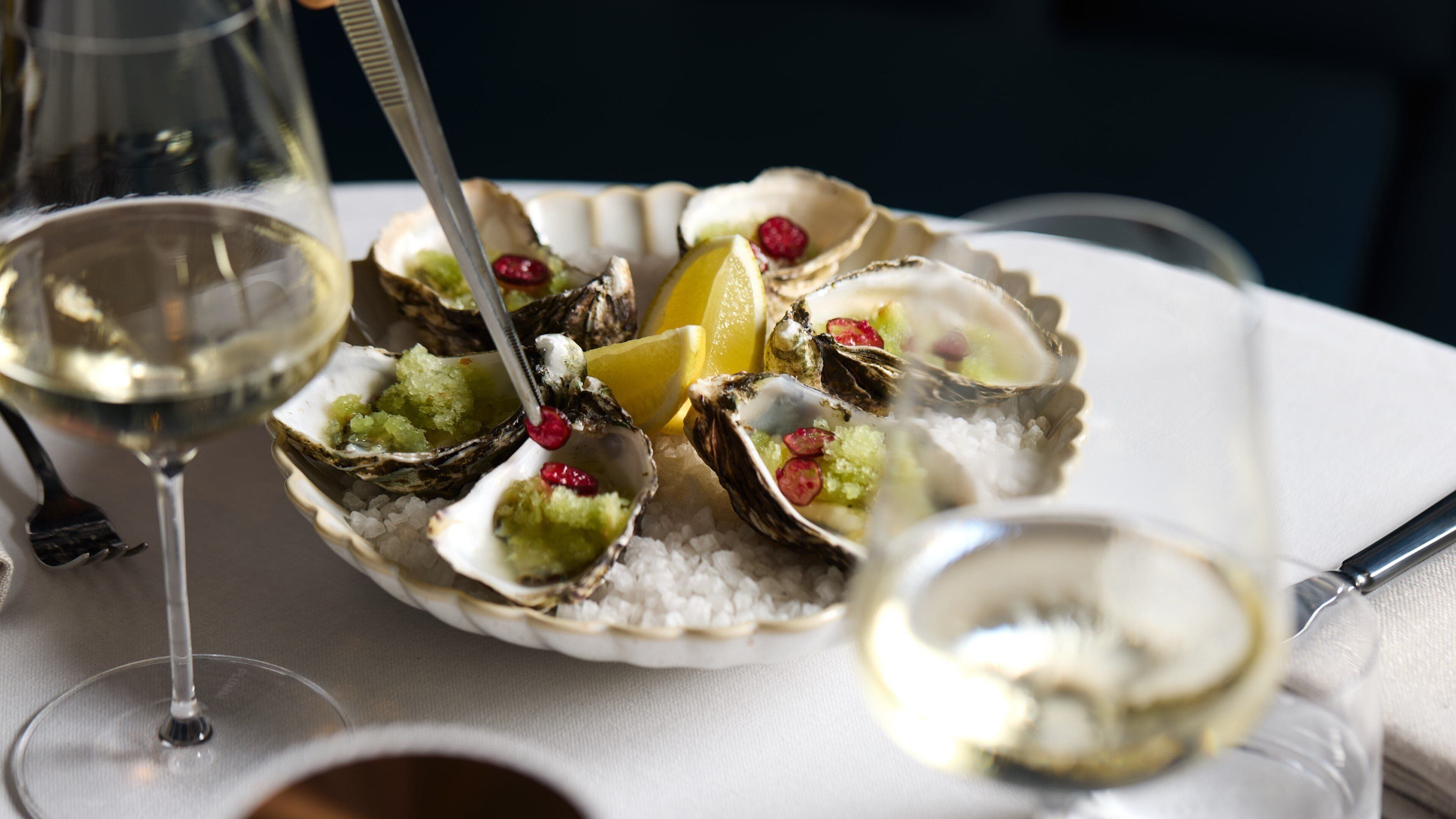 A plate of oysters with lemon wedges and a fork, accompanied by a glass of white wine on a table.
