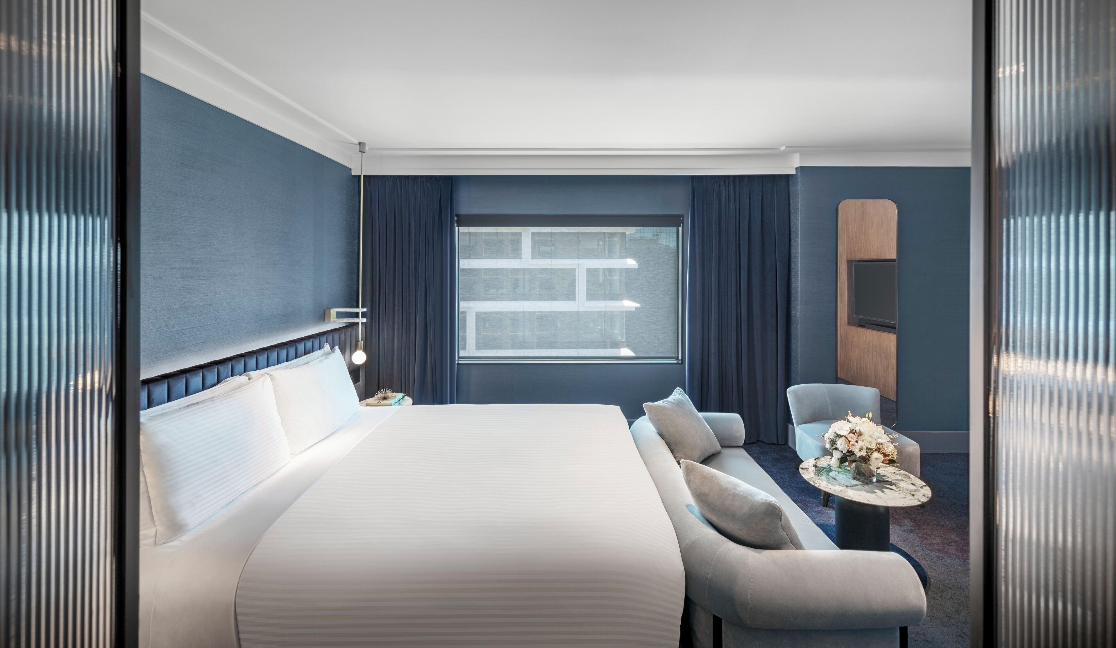 Una habitación de hotel moderna con una cama grande, un área de descanso con una mesita pequeña y un ventanal amplio.