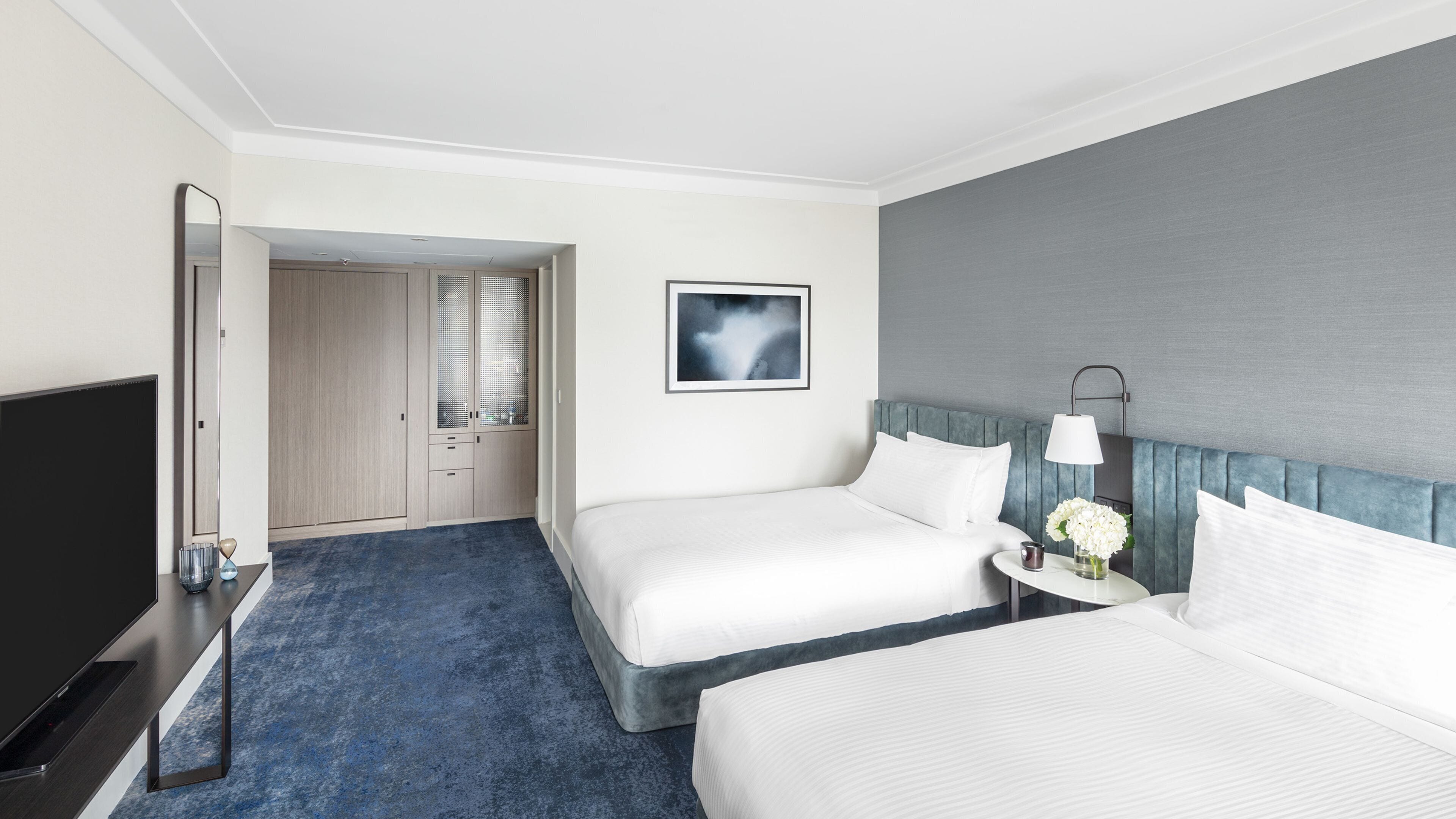 A hotel room with two beds, a TV, a bedside table with a lamp, and a picture on the wall.