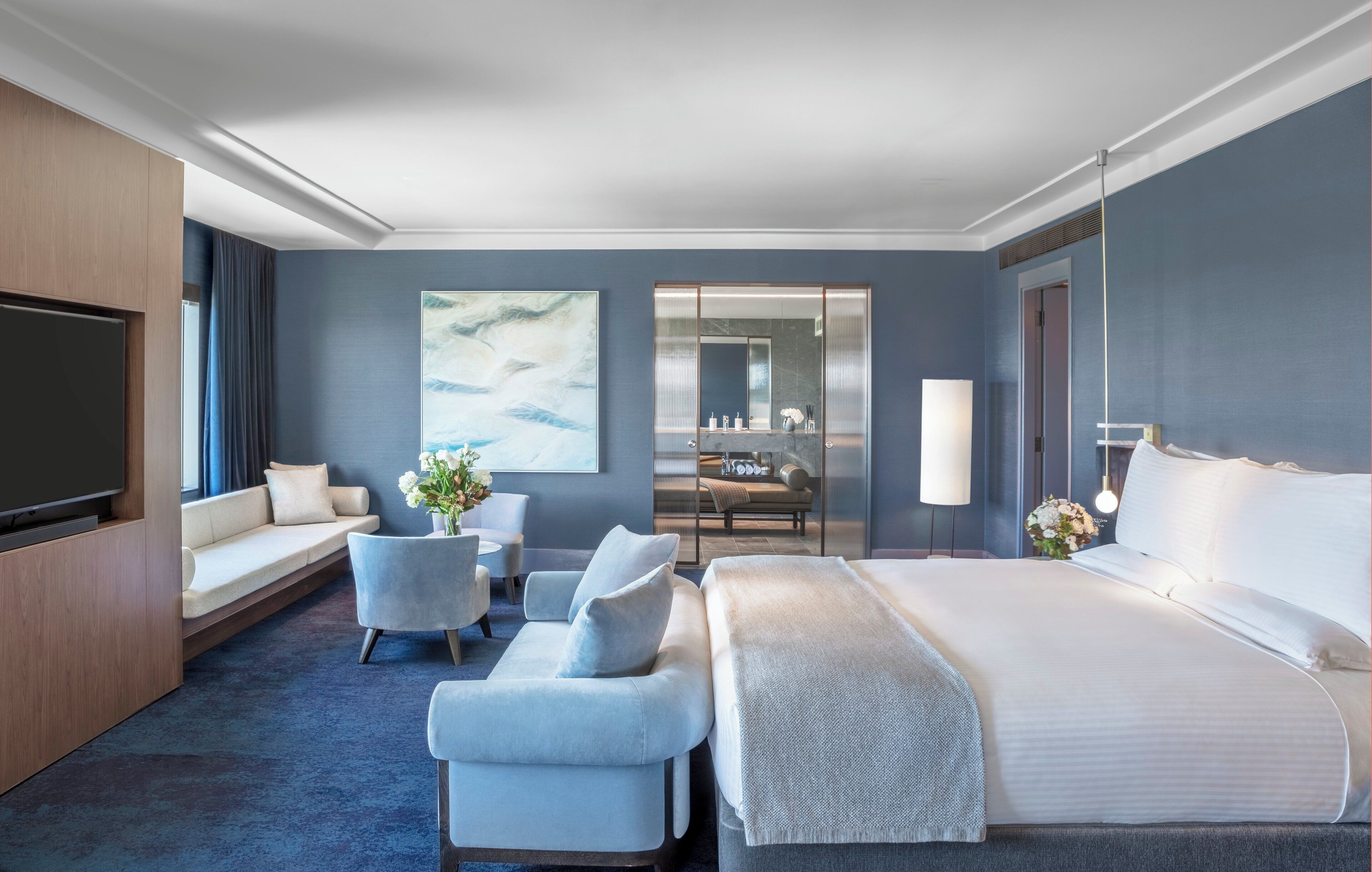 A modern hotel room with a large bed, a seating area with a sofa and armchairs, a flat-screen TV, a dining table with chairs, and a large mirror.