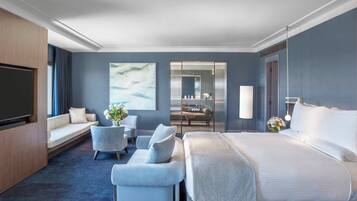 A modern hotel room with a large bed, a seating area with a sofa and armchairs, a flat-screen TV, a dining table with chairs, and a large mirror.