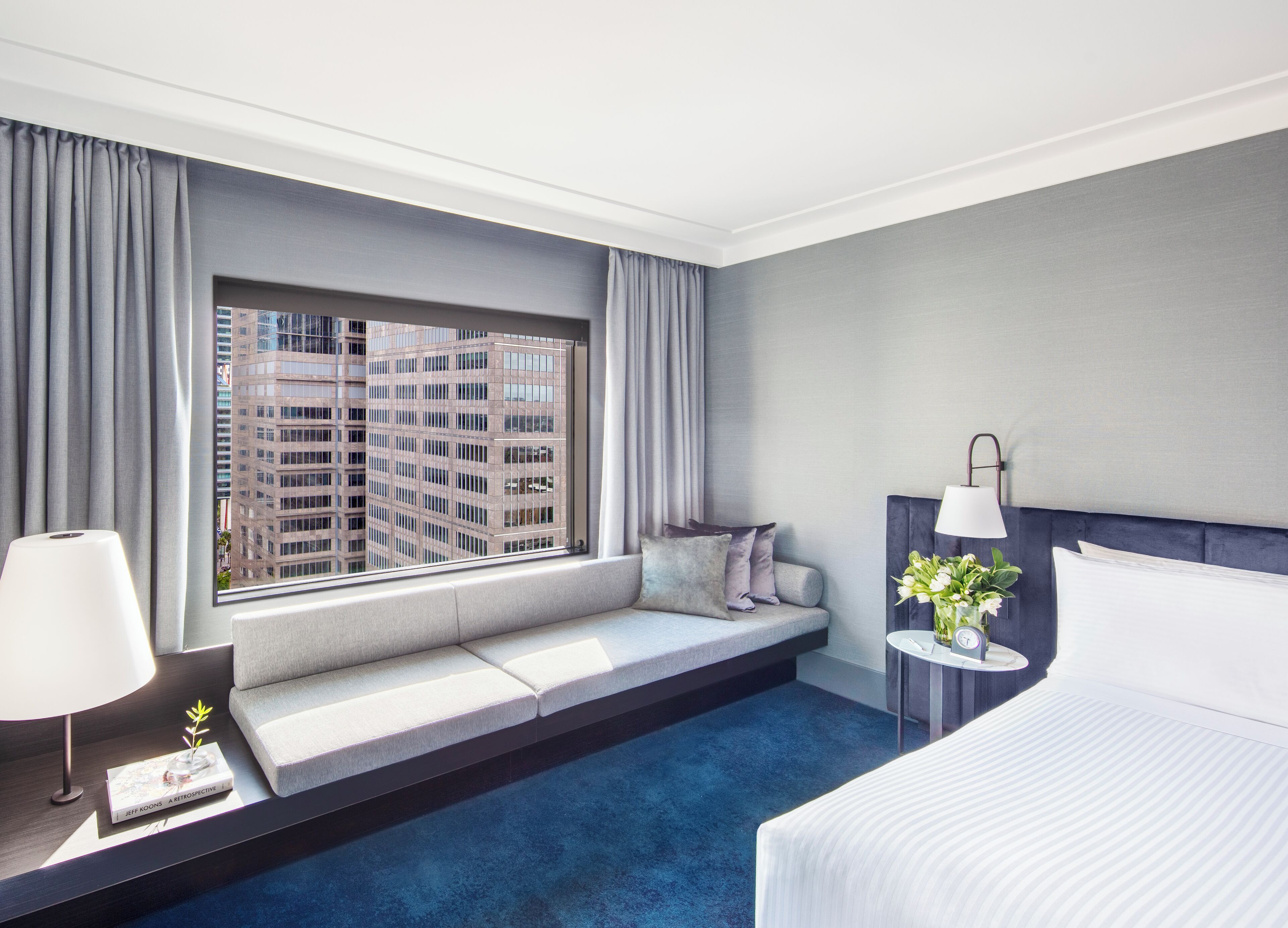 A hotel room with a large window, a sofa bed, a bedside table with a lamp, and a bed.