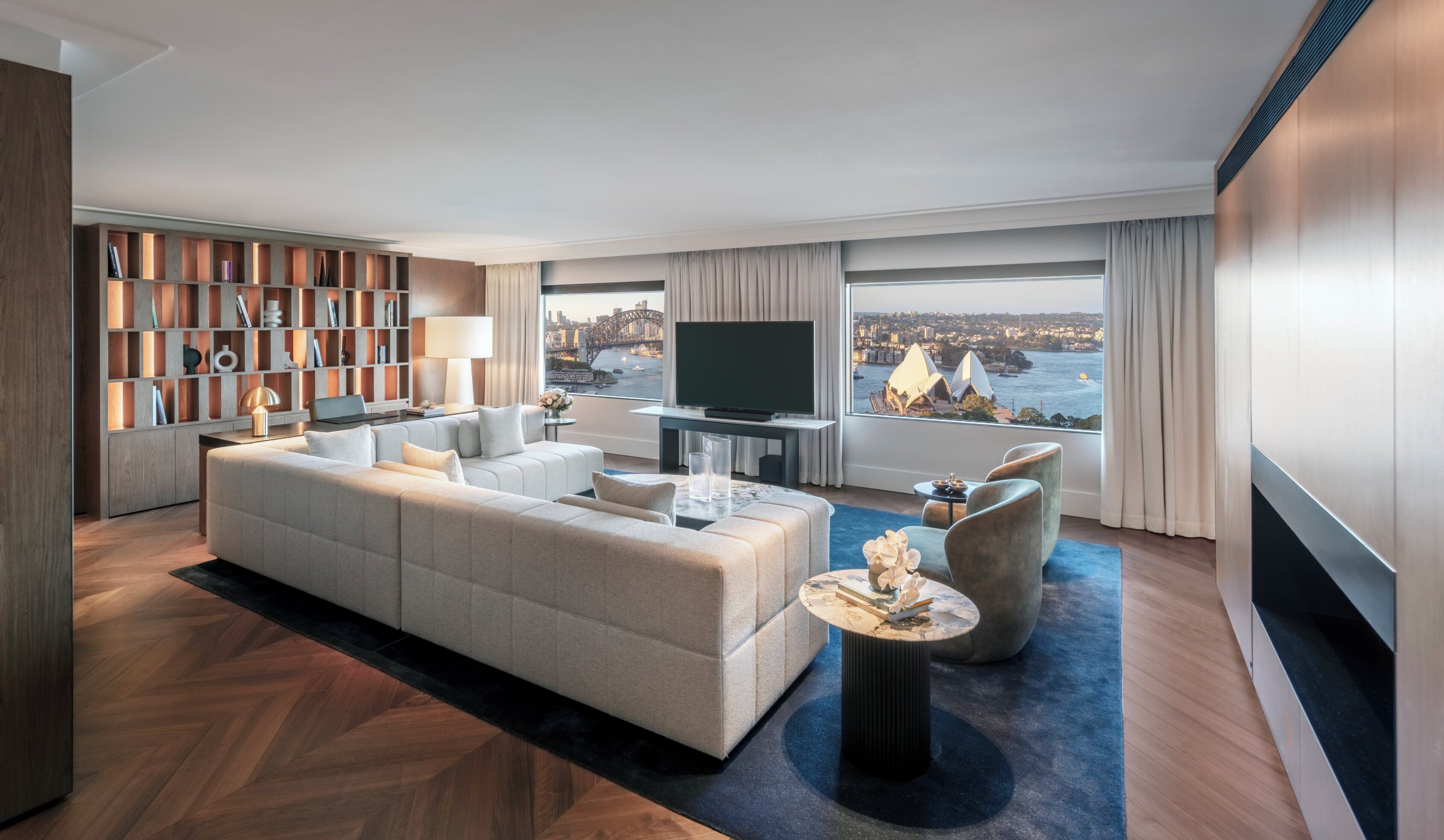 Ein modernes Wohnzimmer mit einem großen Sofa, einem Couchtisch und einem Fernseher. Ein Bücherregal ist mit Büchern und Dekoration gefüllt. Große Fenster bieten Blick auf eine Stadtlandschaft und eine Brücke.