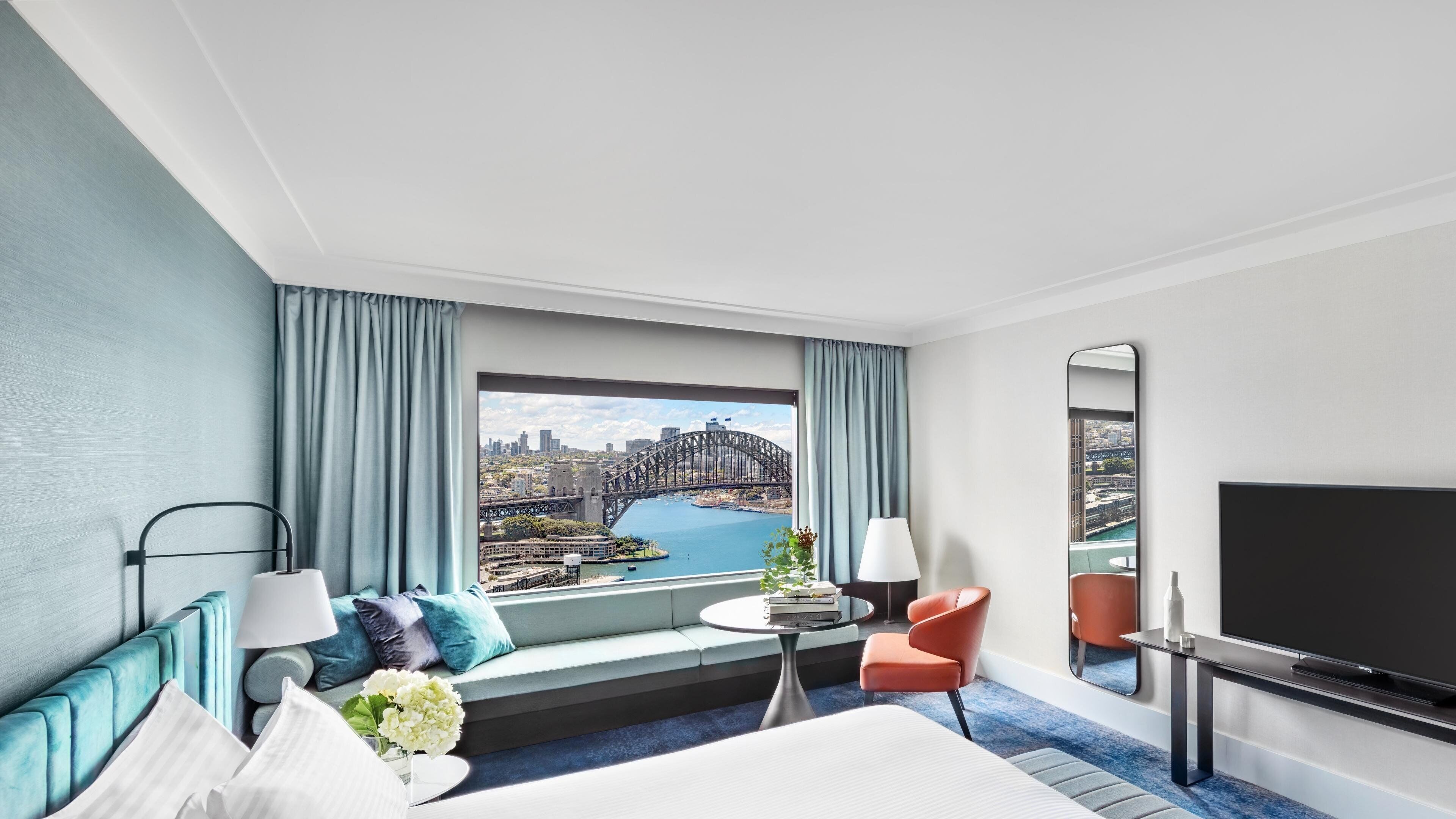 A hotel room with a large bed, a TV, a seating area with a view of a cityscape and a bridge.