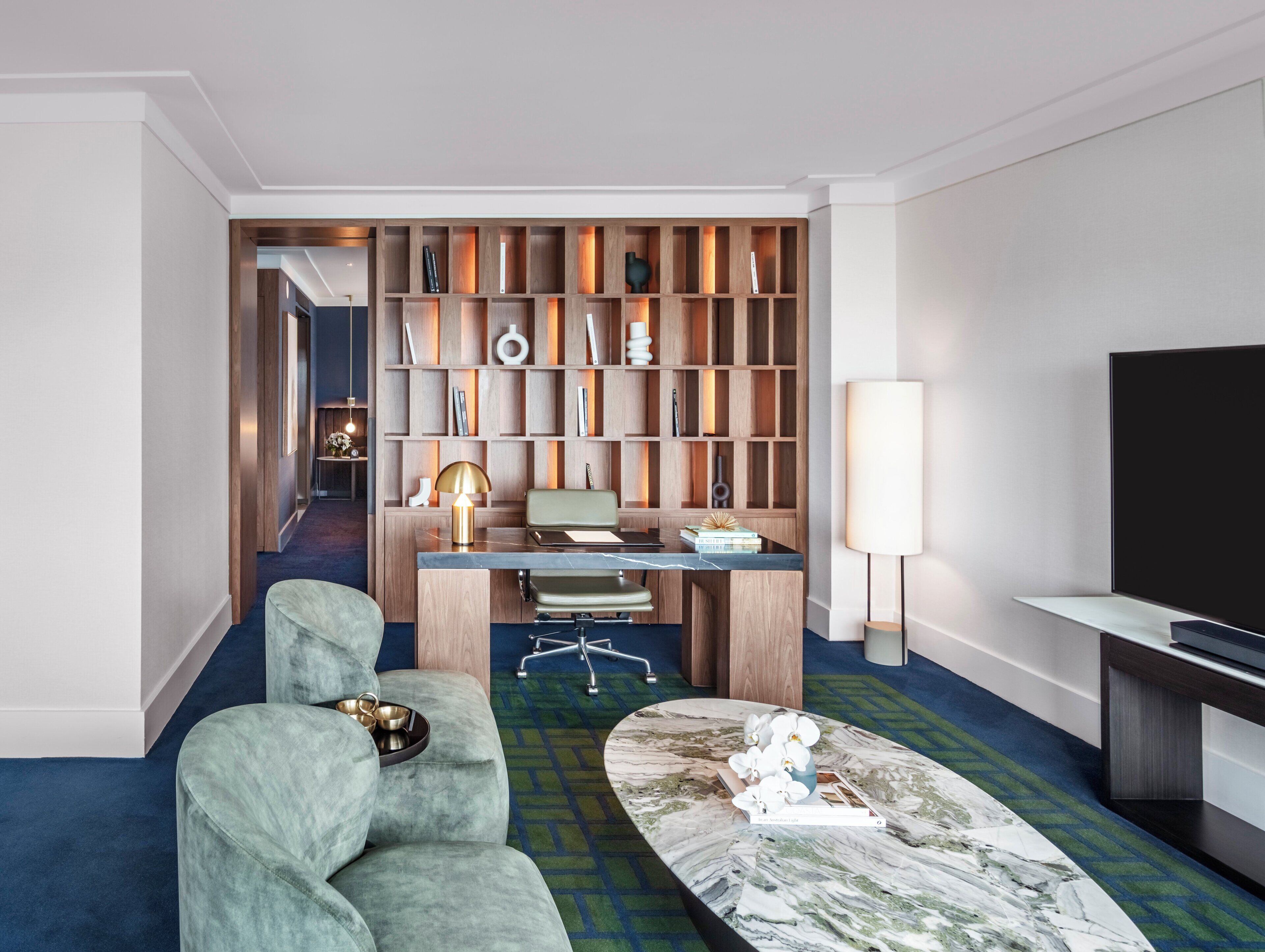 A modern hotel room with a seating area, a desk with a lamp, a television, and a bookshelf.