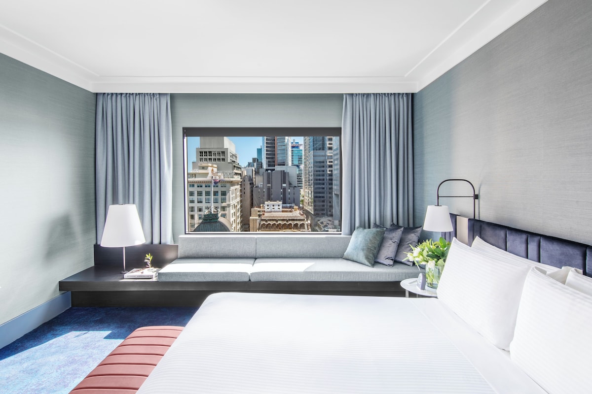 A hotel room with a large bed, a seating area with a view of the city, and a nightstand with a lamp.