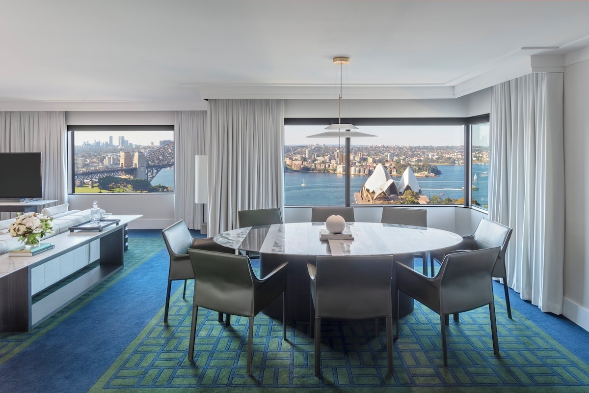A hotel room with a large round table, modern chairs, a flat-screen TV, a sofa, and a view of a cityscape and harbor.
