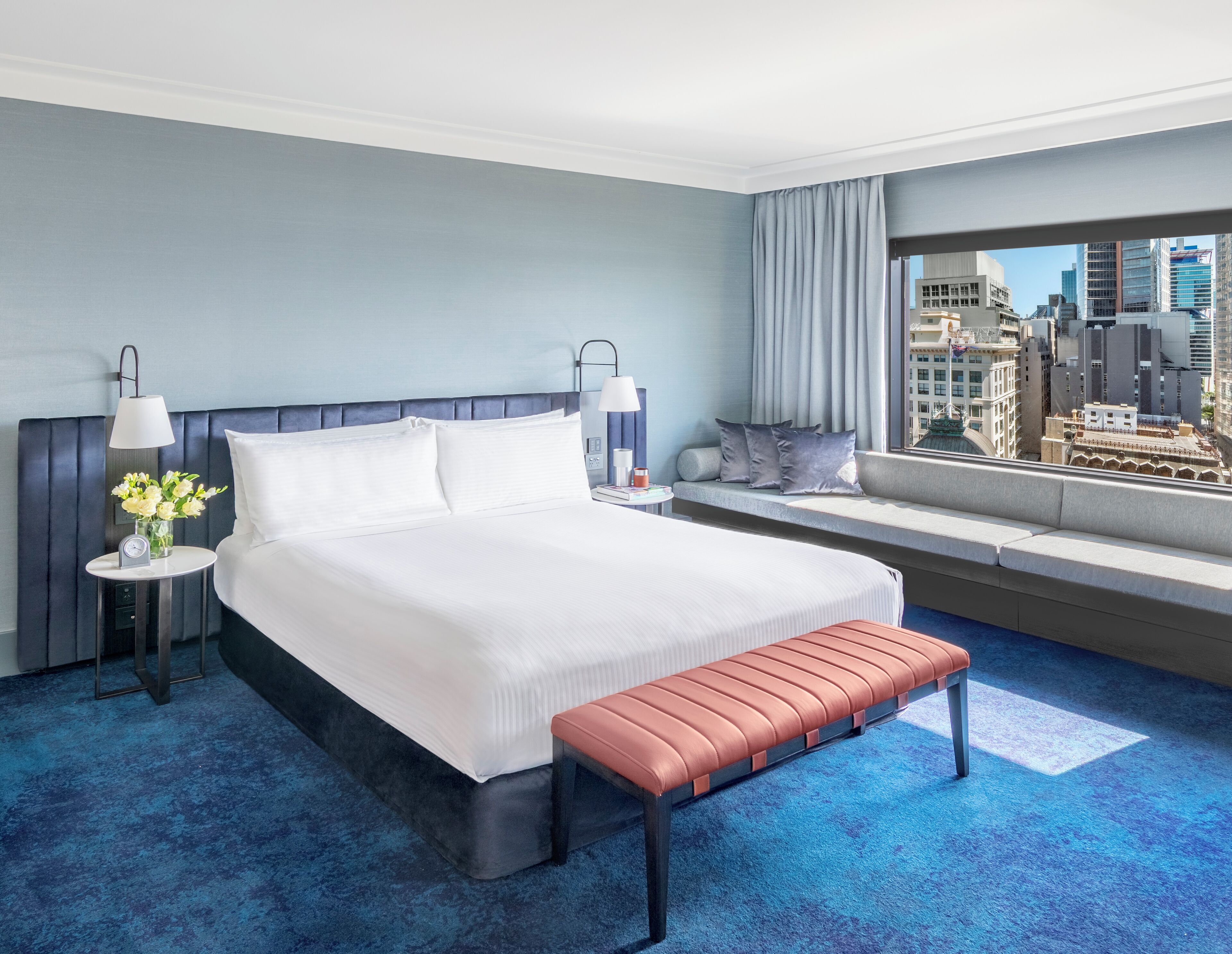 Una habitación de hotel con una cama grande, un banco, una mesita de noche con una lámpara, una ventana con vistas a la ciudad y vistas urbanas desde la ventana.
