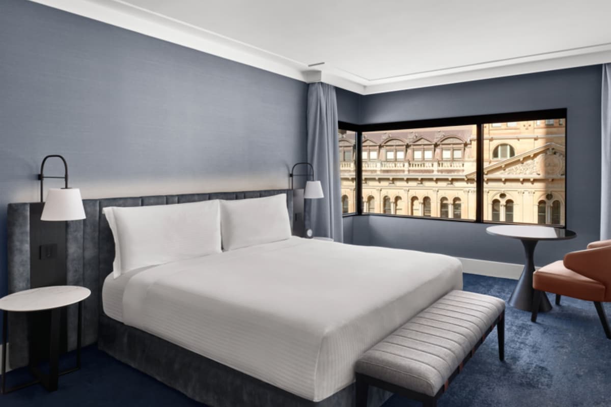 A modern hotel room with a large bed, a small table, a chair, and a view of a historic building.