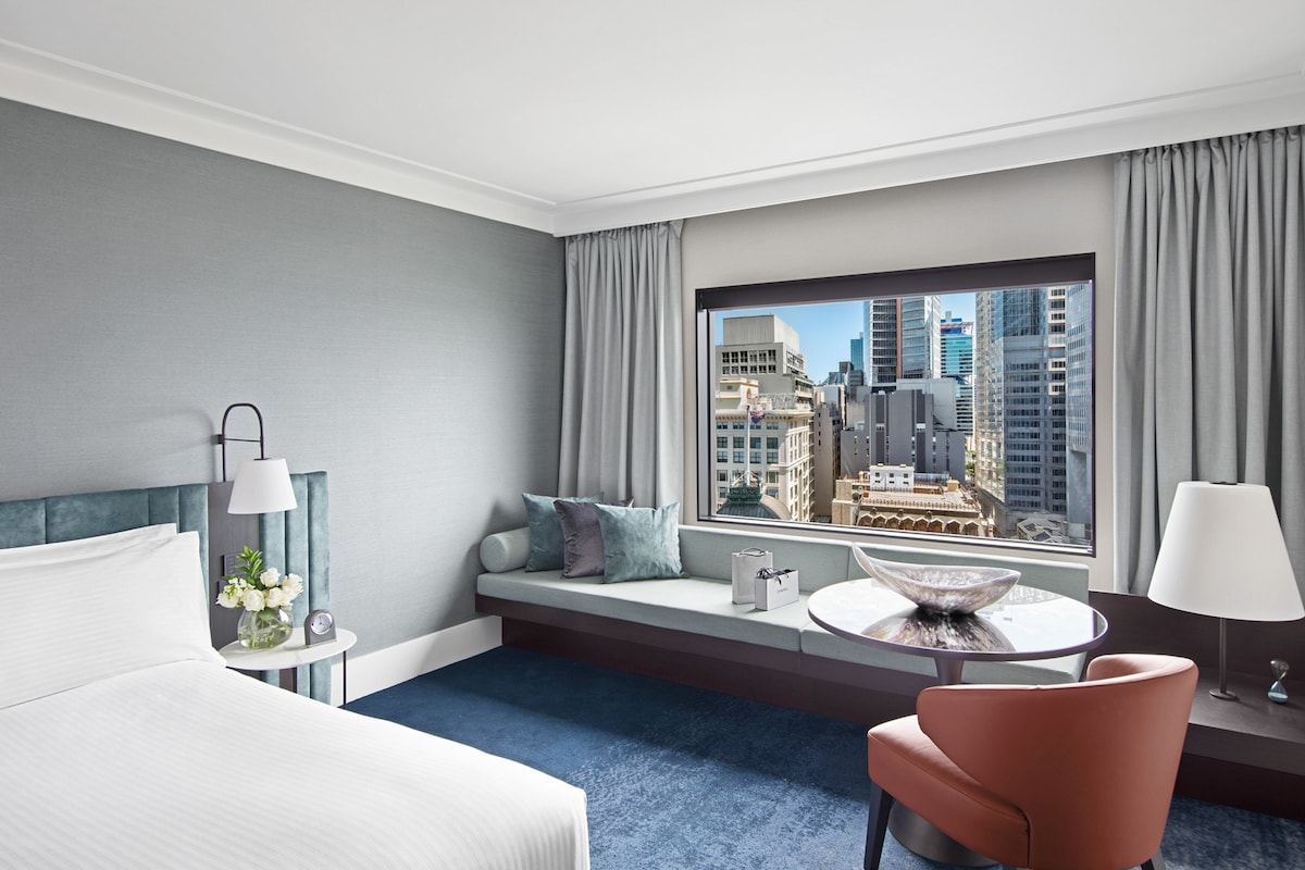A hotel room with a bed, a seating area with a view of the city, a round table, and a chair.