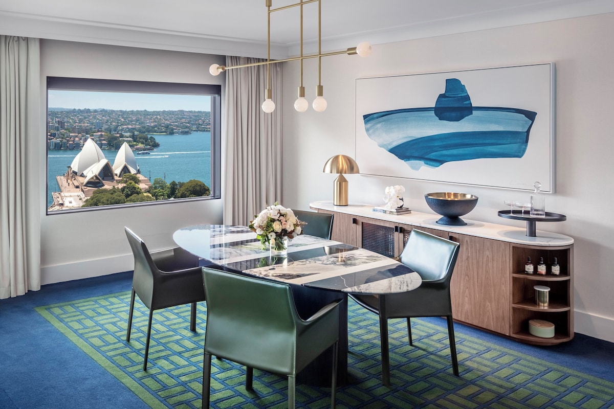 A modern hotel room with a dining area featuring a round table, chairs, and a view of the Sydney Opera House.