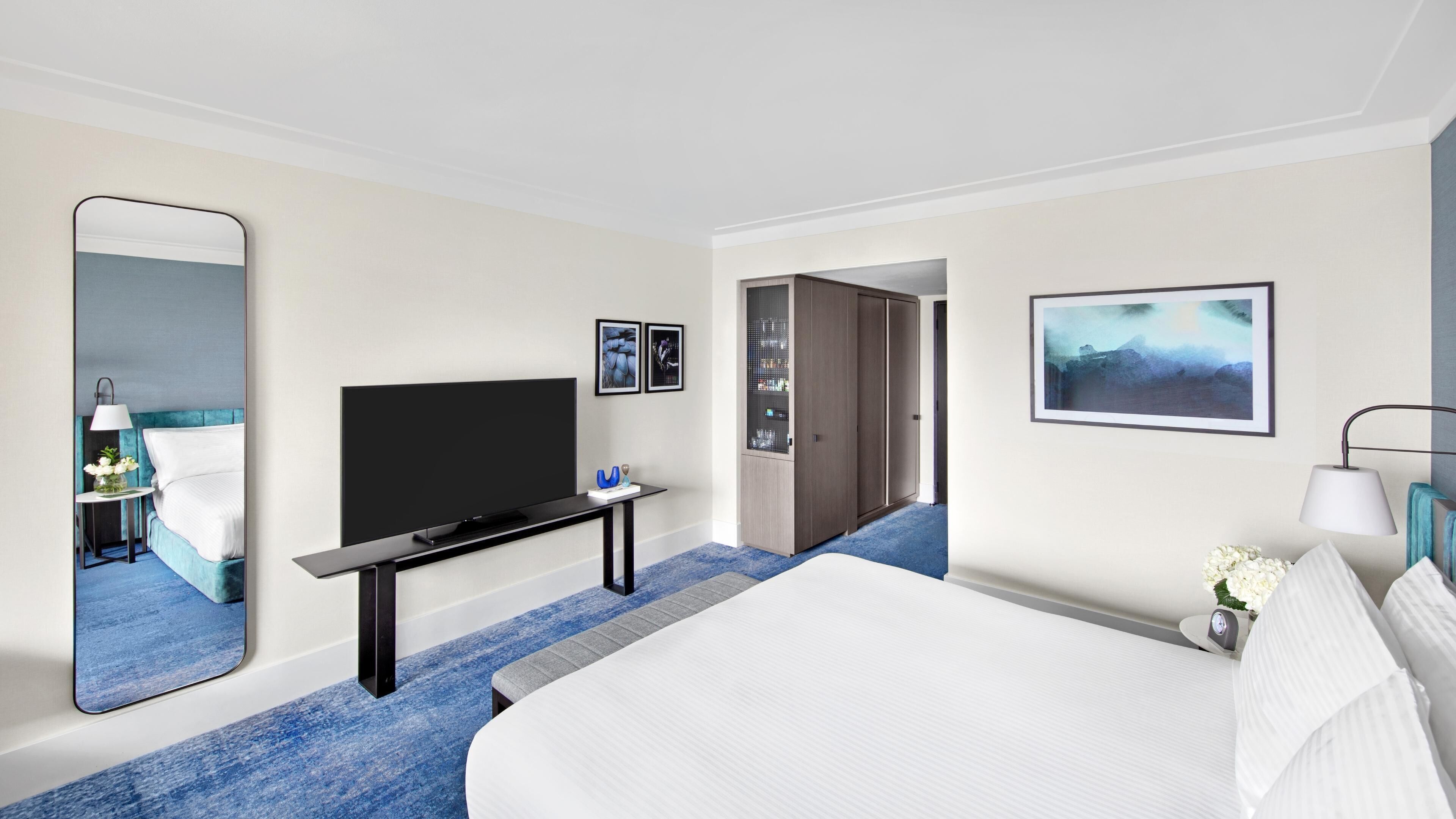 Una habitación de hotel con una cama, un televisor en un soporte, un espejo y una puerta que da a otra habitación.
