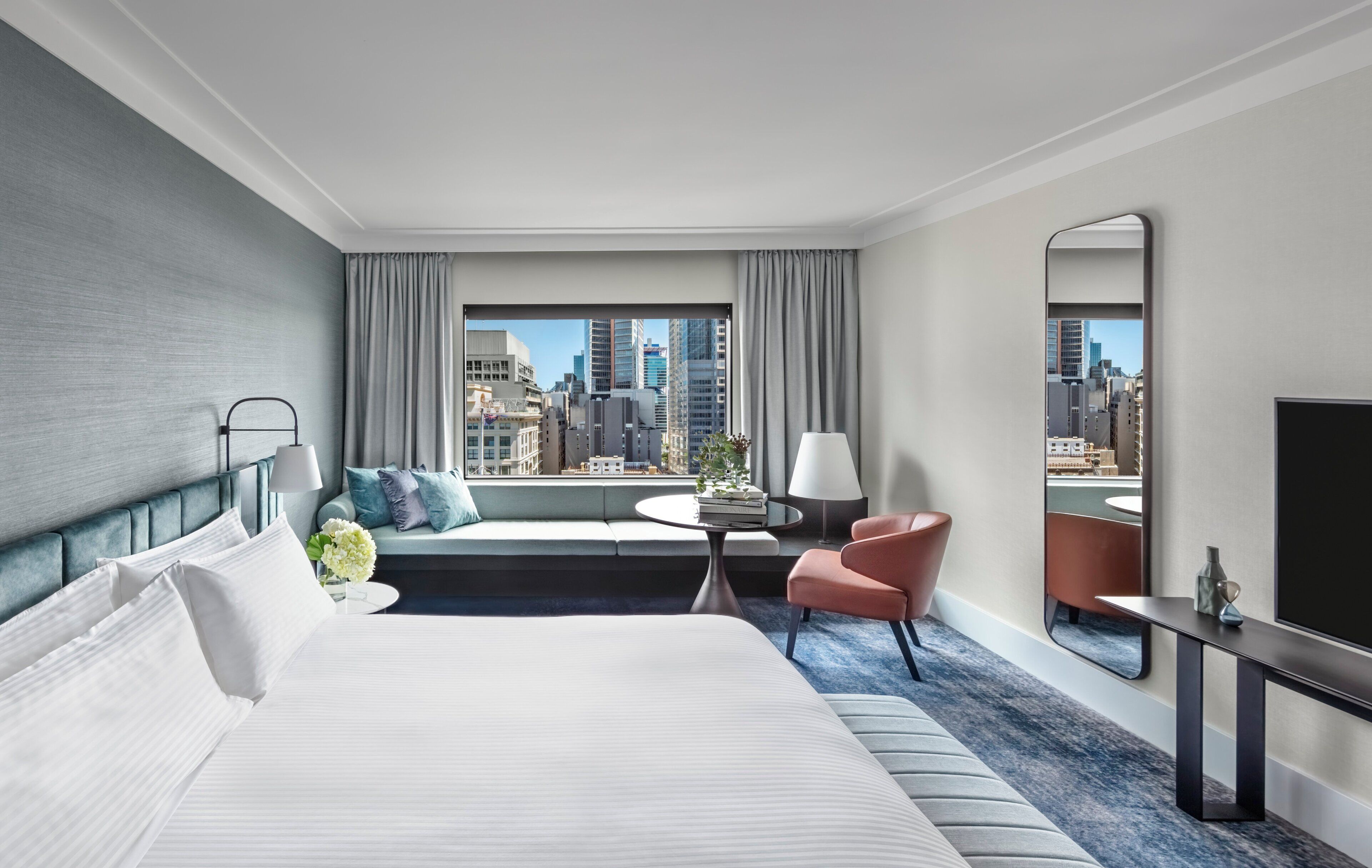 A hotel room with a large bed, a sofa, a small table, a chair, a TV, and a city view.