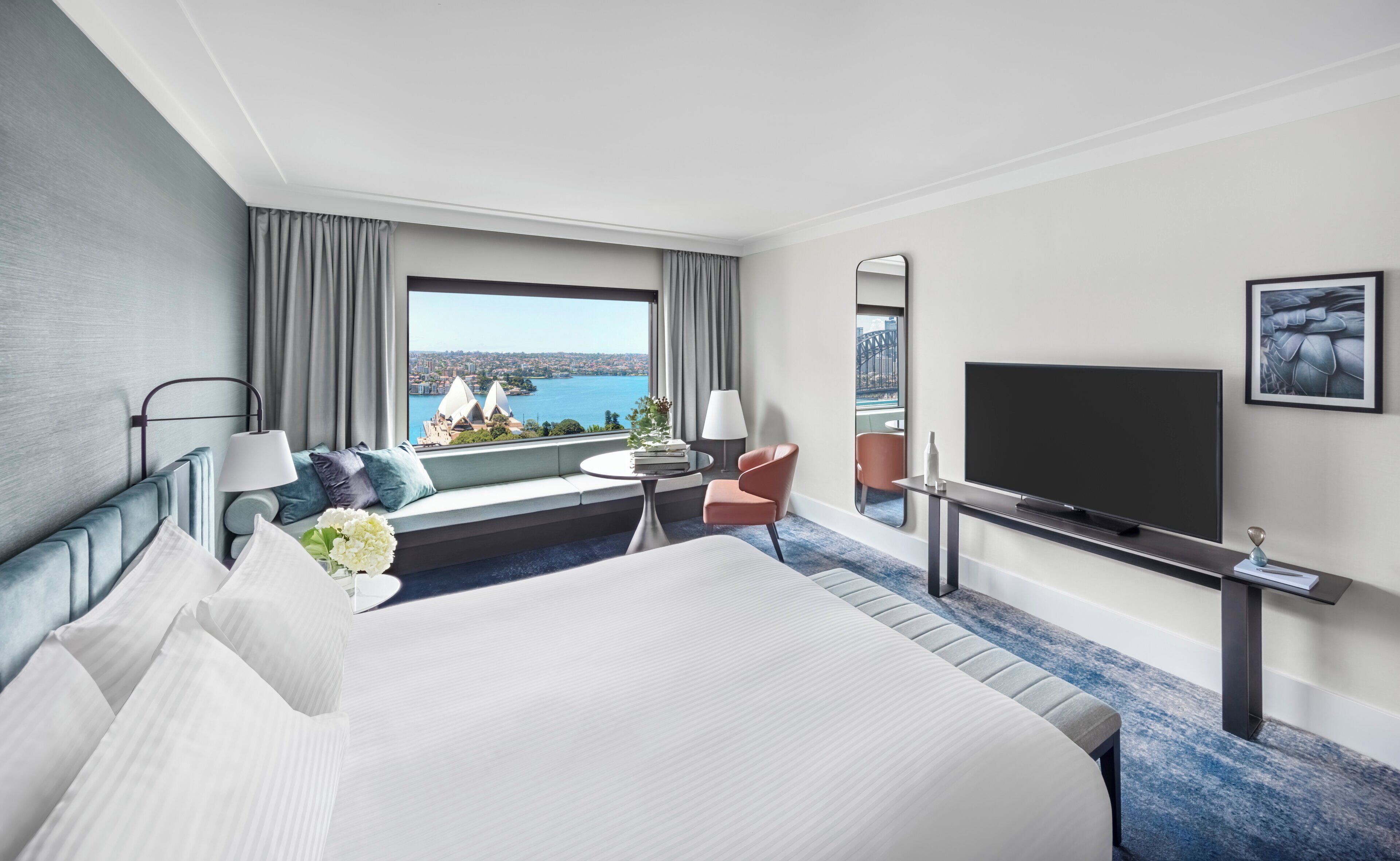 A hotel room with a large bed, a TV, a seating area with a view of a cityscape and water, and a window with curtains.