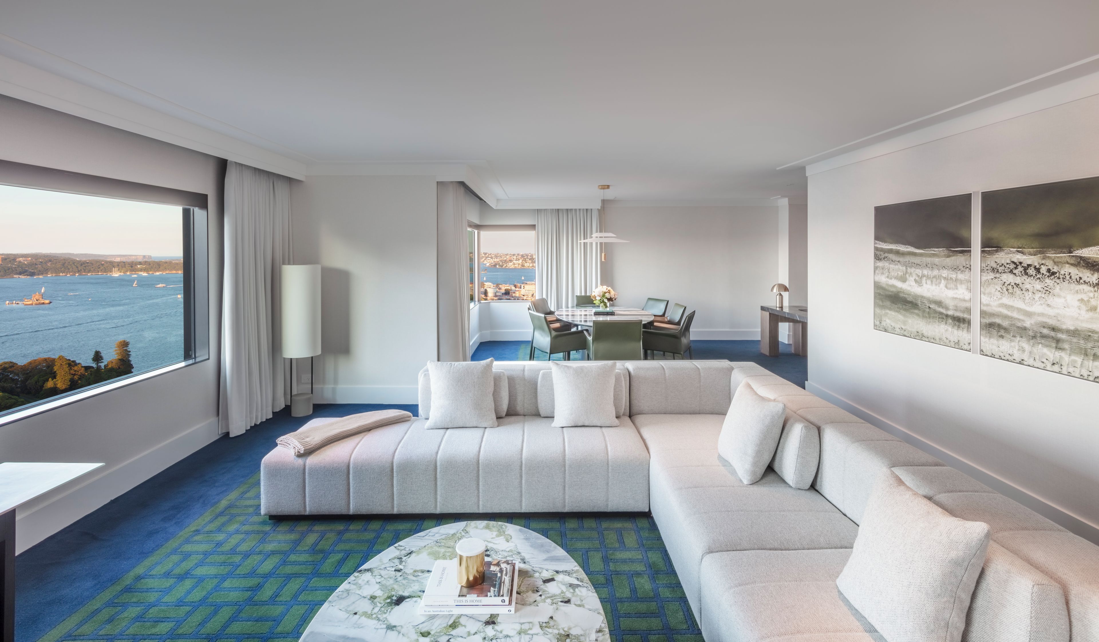 A modern hotel room with a large window offering a view of the sea, a comfortable sofa, a dining area, and a desk with a laptop.